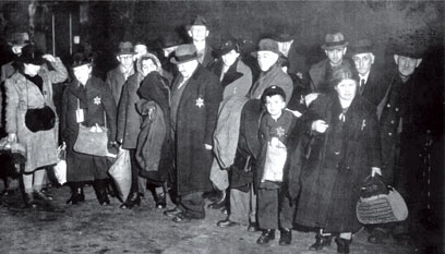 18 persons, including a child, standing close together with bags and sacks in their hands, the majority with hats on their heads. They bear the “Star of David” on their left breast.