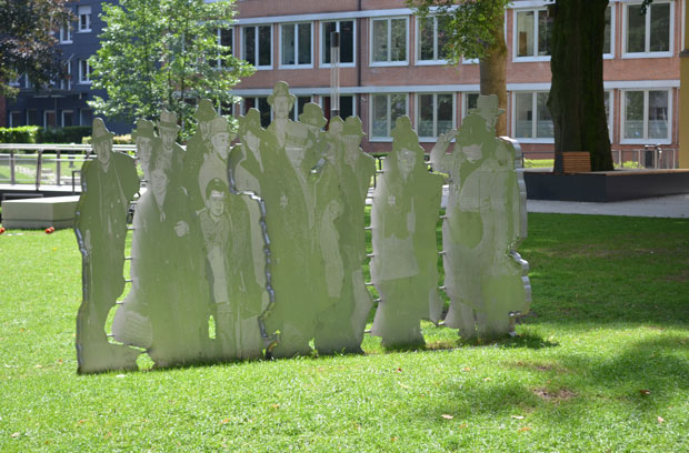 Steel sculpture of people, on a lawn with paths and buildings nearby.