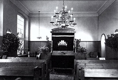 Black-and-white photograph with benches to the left and right; above the centre aisle, a small chandelier; at the front, a Torah shrine.